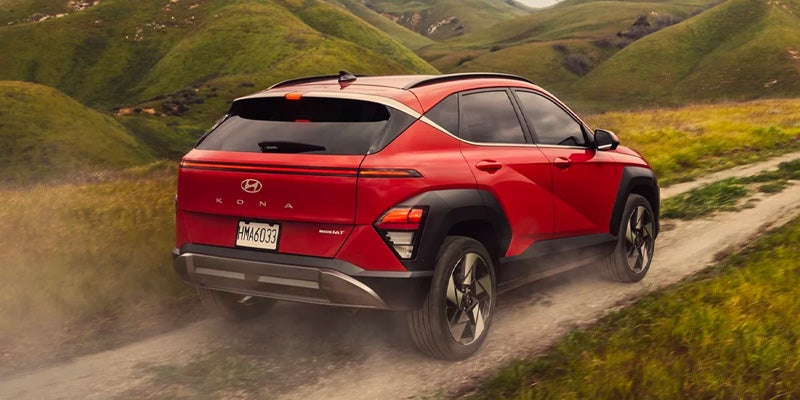 A red 2024 Hyundai KONA driving along a dirt road surrounded by greenery.